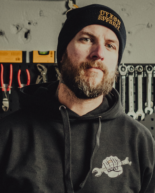 Man wearing a black hoodie and beanie with 'Dyxon Apparel' branding in a workshop setting.